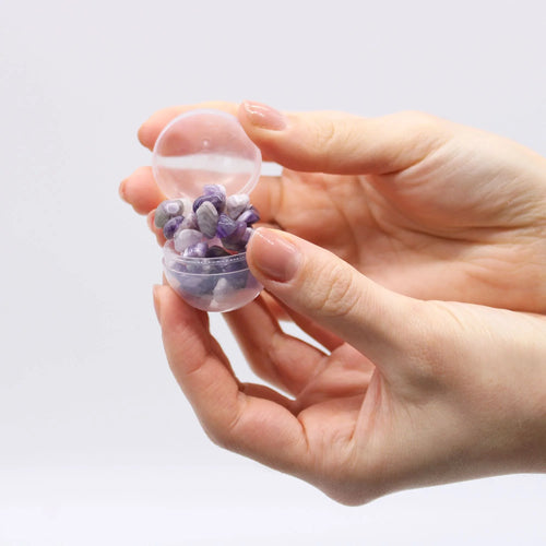 Hand holding a small plastic capsule with tumbled purple crystals inside